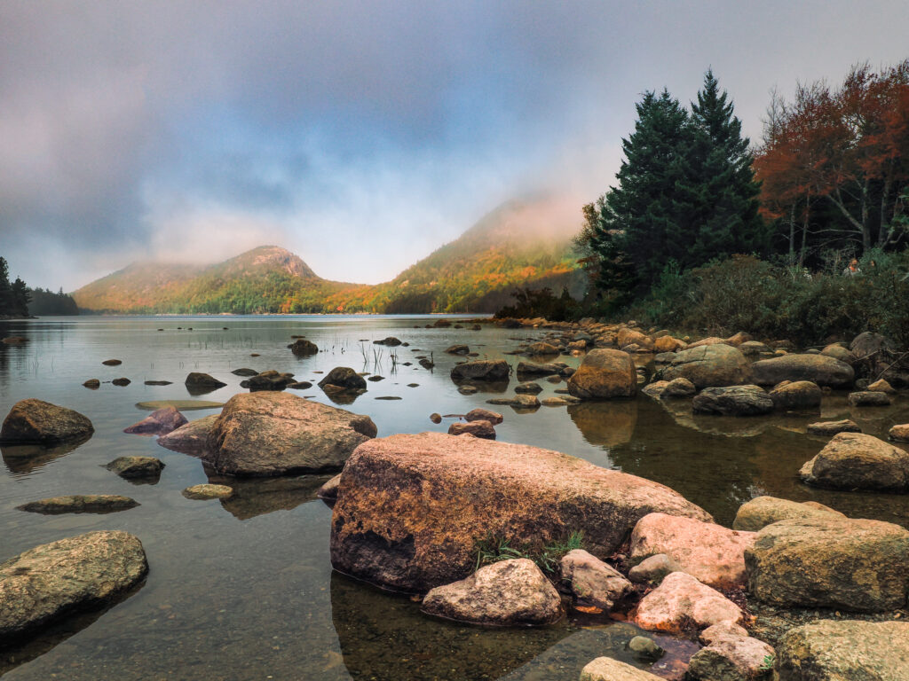 The Bubbles, two famous mountains near Jordan Pond in Acadia National Park, are cloaked in morning fog at sunrise. Large pink granite rocks sit in the lake in the foreground.