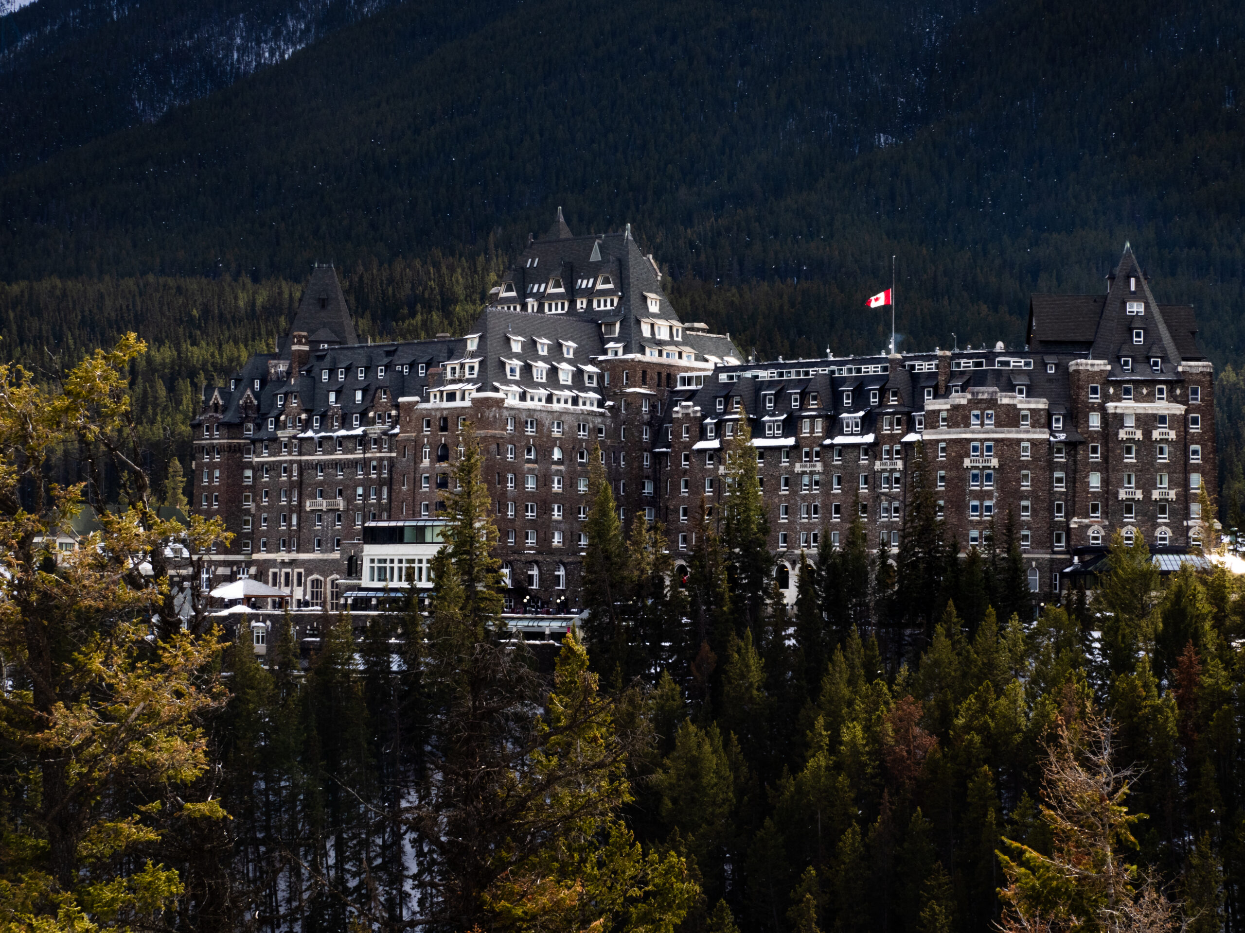 Fairmont Banff Springs, a large castle hotel, from afar, with trees in the foreground and mountains in the background.