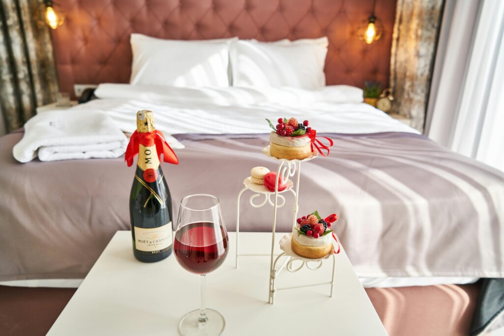 Brut wine, macarons, and French pastry decorated with ribbon on table in front of hotel bed