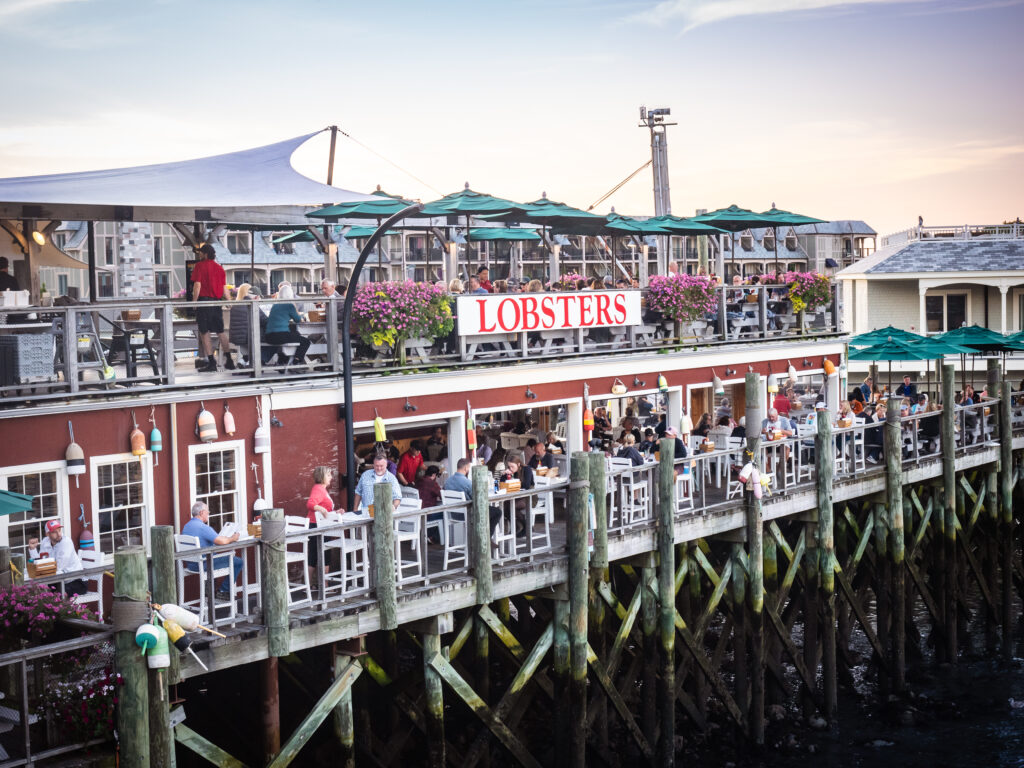 The outdoor patio at Stewmans Lobster Pound in Bar Harbor, Maine, filled with people standing and sitting during a sunset meal.